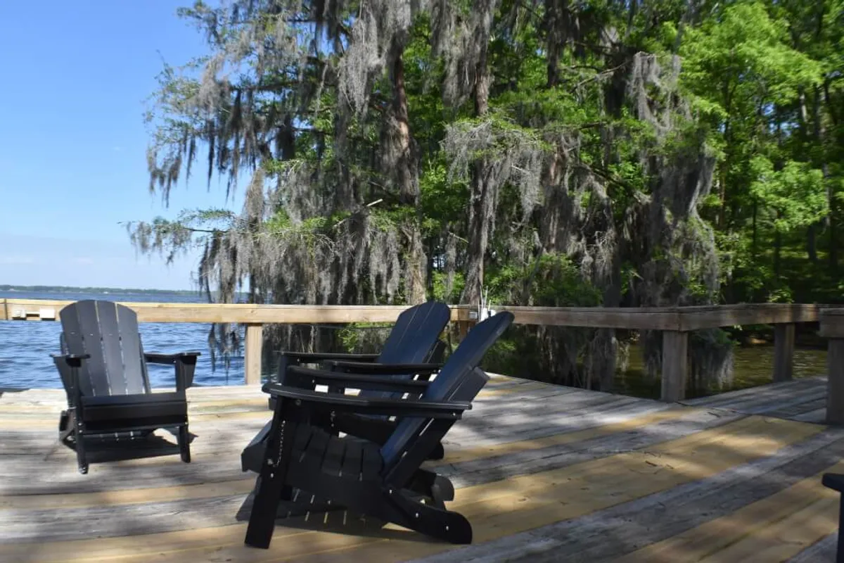 A wooden deck outfitted with Adirondack chairs overlooks the waterfront, framed by Spanish moss hanging from nearby trees. The scene evokes serenity, with cool breezes and a perfect view for stargazing, sunrise, or simply soaking in the lakeside ambiance.