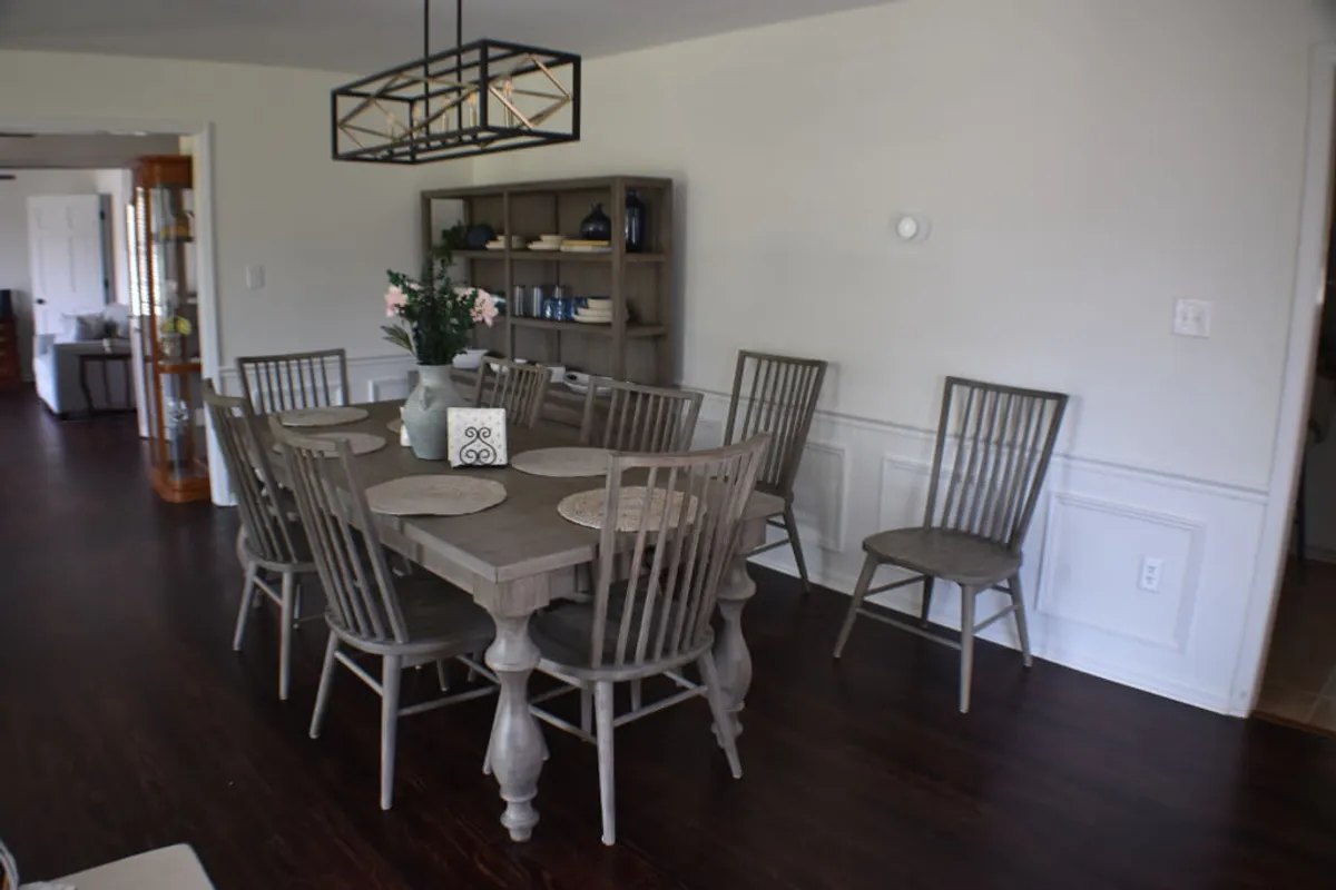A bright, inviting dining room with a wooden dining table set beneath a chandelier, a bookshelf adorned with decor, and a centerpiece floral arrangement. Natural light pours in from the window, illuminating a space perfect for family meals and celebrations.