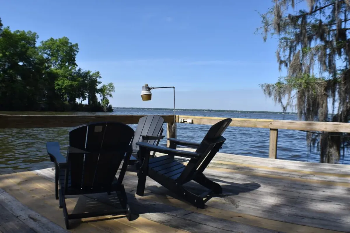 This exterior image shows two black Adirondack chairs positioned right by the lakefront, with a lamp nearby and tranquil water in the background. The setting is peaceful, ideal for relaxing, reading, or simply admiring the lakeside view as the sun sets.