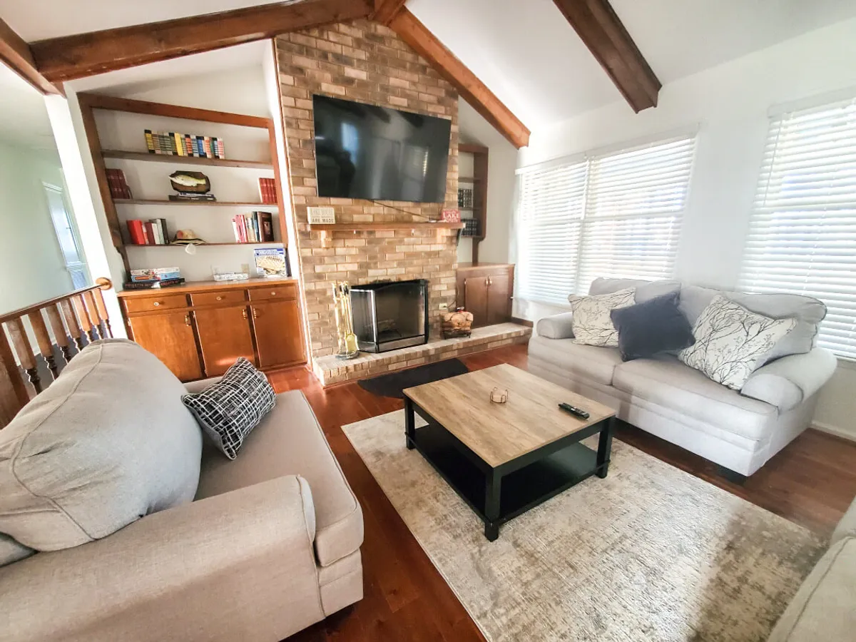 A welcoming living room featuring a brick fireplace as the centerpiece, large TV, a comfortable coffee table, and plush sofas arranged for conversation. The setting exudes comfort, with warm lighting and inviting decor perfect for family relaxation or storytelling.