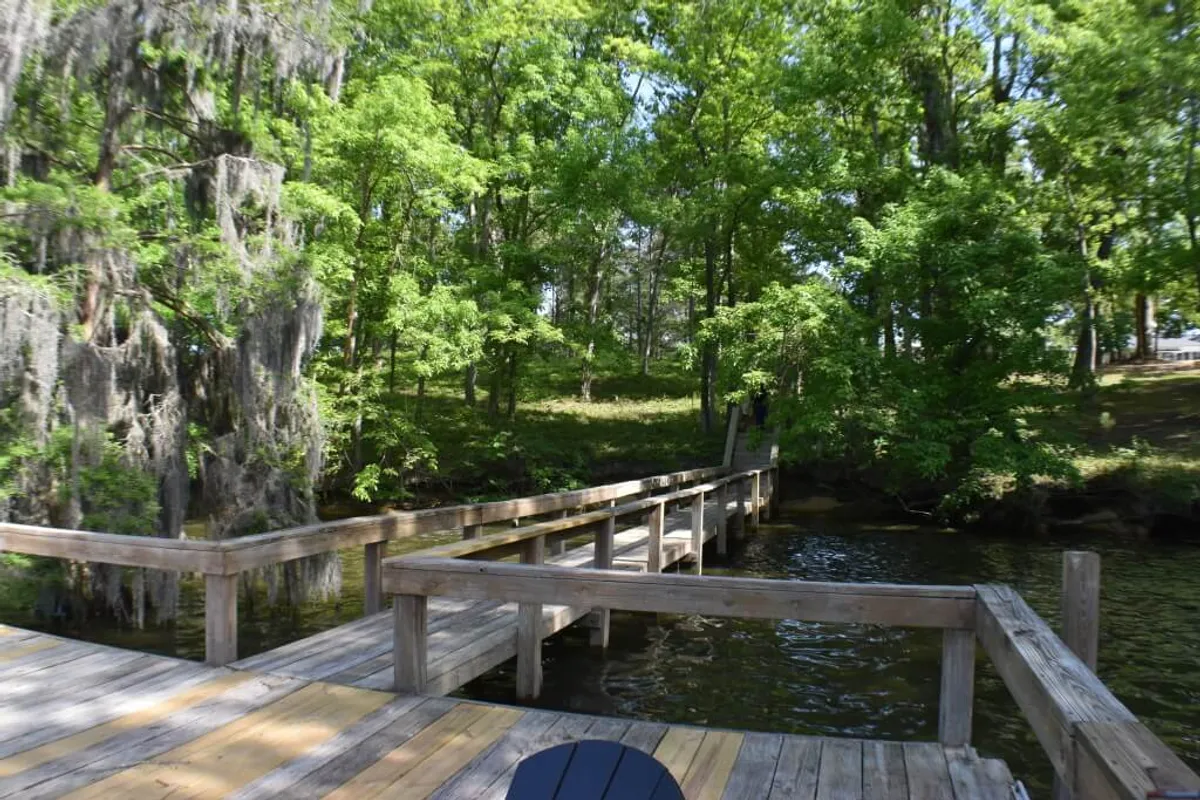A wooden dock extends over calm lake waters, surrounded by lush greenery and shaded by trees. The dock is clearly accessible from the backyard, inviting visitors to enjoy fishing, boating, or simply relaxing at the water's edge.