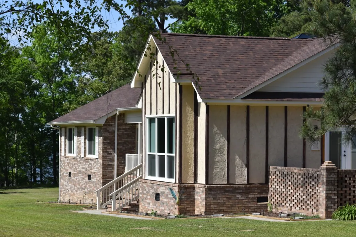 The image shows the welcoming exterior of the lakeside home, highlighting the large front windows, brickwork details, and textured wall panels. The porch is bathed in sunset light, suggesting a tranquil atmosphere perfect for relaxing after a day of adventure.