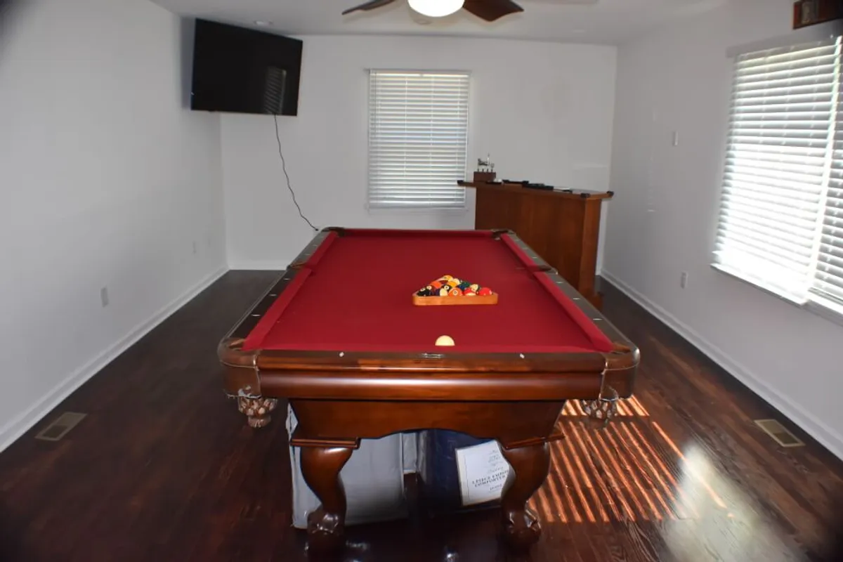 This interior image features a spacious game room with a pool table at its center, a television mounted nearby, and a bar area in the background. The scene is set up for fun and friendly competition, with ample space for guests to gather and enjoy social games.