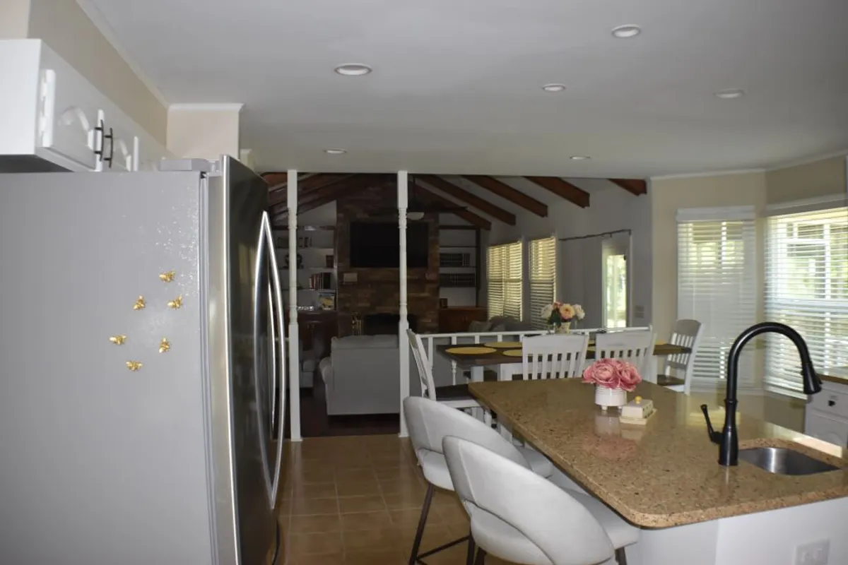 A bright kitchen and dining area, complete with a spacious granite island, white cabinetry, refrigerator, and a wooden dining table. The layout invites both cooking and casual dining, ideal for communal feasts.