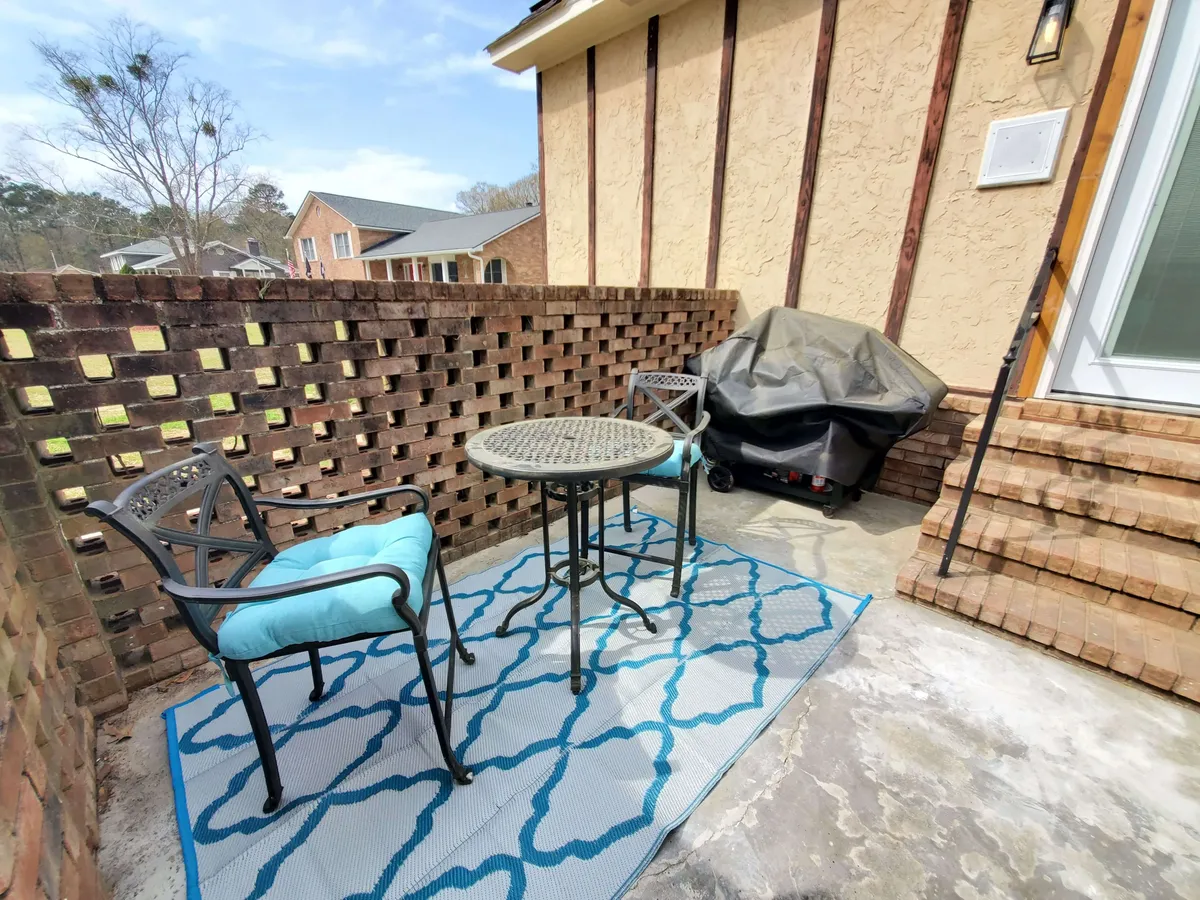 A cozy exterior patio scene featuring a bistro table and two chairs on a decorative rug against a brick wall. It's an intimate spot for morning coffee, afternoon reading, or unwinding at sunset, with hints of garden life visible.