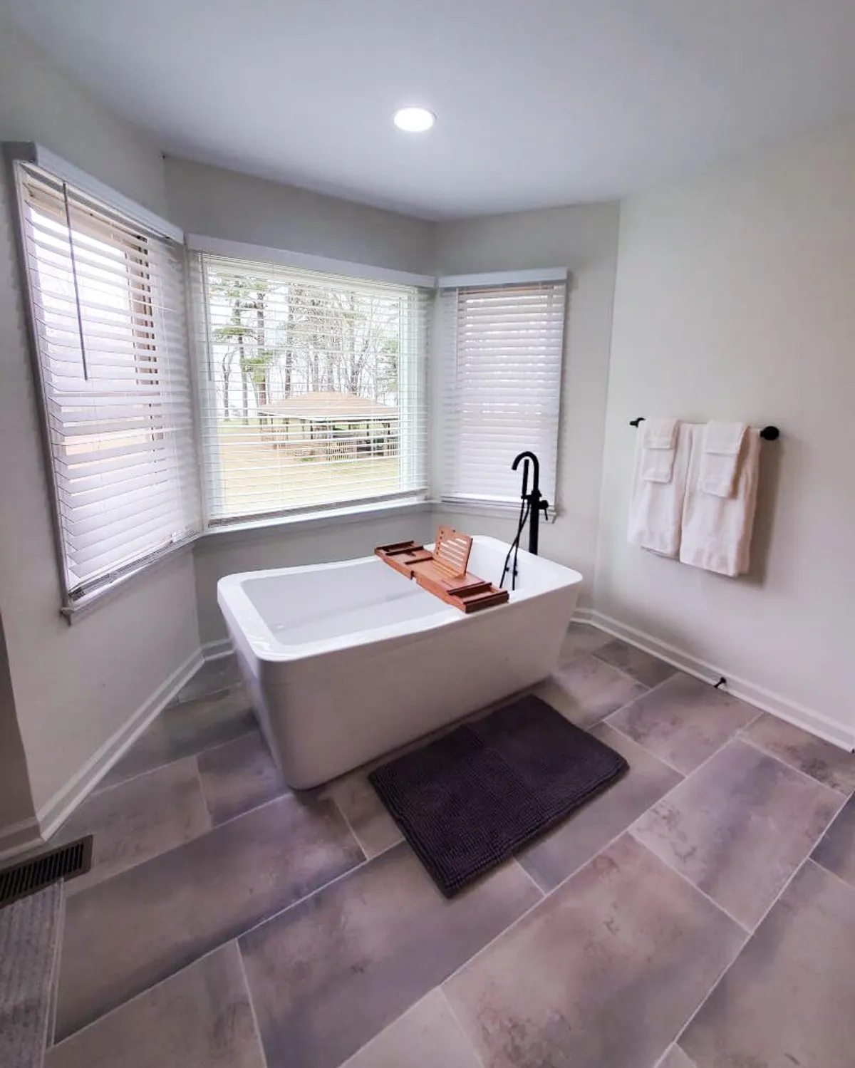 A spa-like bathroom featuring a freestanding bathtub with a wooden bath tray, large windows that frame the landscape, and plush towels nearby. The atmosphere is tranquil and luxurious, suggesting a perfect spot for relaxation with a view.